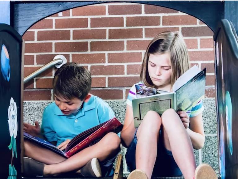 Two children sitting on a bench reading books.